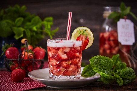 Erdbeer Bowle - Im Vordergrund ist ein Glas mit Erdbeerbowle auf einem weiß-goldenen Teller dargestellt. Dieses ist mit einer Erdbeere, einer Zitrone und einem Strohhalm versehen. Melisse und weitere Erdbeeren im Hintergrund verzieren den Cocktail. (Foto: VrK/Franz Gleiß - Nicht zur freien Verwendung)