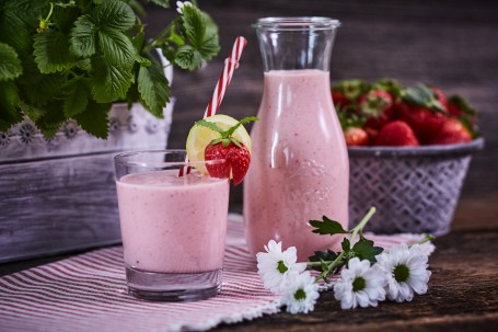 Erdbeer Joghurt Drink - Der Erdbeer Joghurt Drink wird in einem kleinen Gläschen auf einem weiß-roten Tischtuch serviert. Er ist durch einen Strohhalm leicht zu trinken. Das Glas ist mit einer Erdbeere und mit einer Zitrone verziert.  Im Hintergrund befinden sich ein gefüllter Krug und Körbe mit Früchten und Erdbeerpflanzen. (Foto: VrK/Franz Gleiß - Nicht zur freien Verwendung)