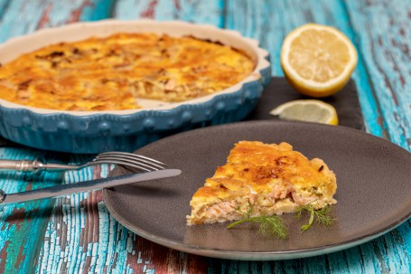 Lachsforellenquiche - Eine blaue Schüssel mit der angeschnitten Quiche steht auf einem blaufarbenen Tisch; dabenben steht ein grauer Teller mit einem dreieckigen Stück der gold/geld gebackenen Quiche, garniert mit Kräuter (Foto: VrK/Achim Mandler Photography - Nicht zur freien Verwendung)