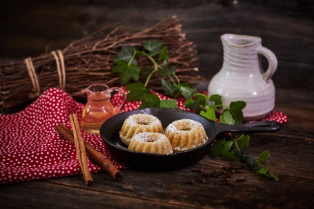 Mostpudding - In einer schwarzen Pfanne befinden sich drei goldbraune Stück Mostpudding. Die Pfanne steht auf einem braunen Holztisch und ist mit einem roten Tischtuch mit weißen Punkten unterlegt. Links neben der Pfanne liegen zwei Zimtstangen. Links hinter der Pfanne befindet sich ein kleiner Glaskrug, der mit Most gefüllt ist. Rechts hinter der Pfanne ist ein weißer Tonkrug zu sehen. Grünes Efeu und ein braunes Bündel Holz dienen der Zierde. (Foto: VrK/Franz Gleiß - Nicht zur freien Verwendung)