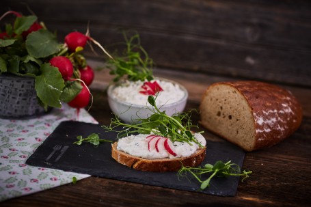 Radieschen-Kräuter Aufstich - Auf einem schwarzen Untersetzer ist ein Brot mit Radieschen-Kräuter-Aufstrich hergerichtet. Das Brot ist mit vier kleinen Radieschen-Scheiben und Kräutern verziert. Auf dem schwarzen Untersetzer befindet sich links ein Symbol für Messer und Gabel. Auf der linken Seite ist ein Tischtuch mit grün-roten Blumen. Darauf befindet sich ein Korb mit Radieschen-Pflanzen. Hinter dem Brot ist eine Schüssel mit Aufstrich, rechts ist ein halber Wecken Brot platziert. (Foto: VrK/Franz Gleiß - Nicht zur freien Verwendung)