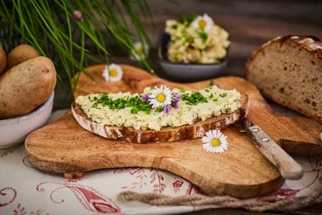 Erdäpfelkas - Auf einem schmetterlingsförmigen Holzuntersetzer ist ein Brot mit Erdäpfelkas zu sehen. Dieser ist mit Schnittlauch, Gänseblümchen und violetten Blüten geschmückt. Rechts unterhalb des Brotes liegt ein Messer. Der Holzuntersetzer befindet sich auf einem weißen Tischtuch mit rotem Blumenmuster. Links ist eine Schale mit Erdäpfeln platziert. Hinter dem Holzschneidbrett ist eine dunkelgraue, kleine Schale mit Erdäpfelkas. Rechts befindet sich ein halber Wecken Brot. Links hinter den Kartoffeln befindet sich Schnittlauch. (Foto: VrK/Franz Gleiß - Nicht zur freien Verwendung)