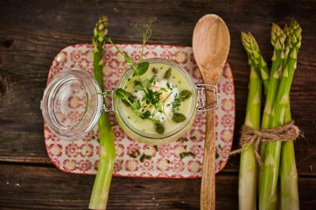 Grüne Spargelcremesuppe - Auf einem rot-gelb gemusterten Teller steht ein Glas, das mit Spargelcremesuppe gefüllt ist. Rechts neben dem Glas liegt ein hölzerner Suppenlöffel. Am rechten Rand des Bildes sowie links auf dem Teller befindet sich grüner Spargel. Das Ganze ist auf einem Holztisch angerichtet. (Foto: VrK/Franz Gleiß - Nicht zur freien Verwendung)