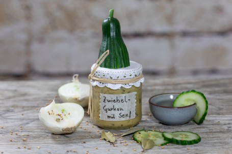 Zwiebel Gurken mit Senf - Auf grauem Hintergrund steht ein Glas mit hellgelbem INhalt, verschlossen mit einem weißen Papier, verschlossen mit einer Schnur; darauf befindet sich ein Stück einer ganzen Gurke, daneben liegt eine aufgeschnittene weiße Zwiebel und ein blaues Schälchen, garniert mit Gurkenscheiben und Lorbeerblätter (Foto: VrK/Achim Mandler Photography - Nicht zur freien Verwendung)