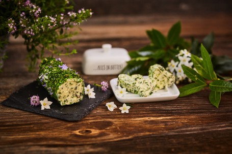 Kräuterbutter - Auf einem Holztisch wird auf einem schwarzen, kleinen Tablett die Kräuterbutter gezeigt. Das Tablett ist mit rosa und weißen Blüten verziert. Die Kräuterbutter ist von kleingeschnittenen Kräutern und Blüten umhüllt. Neben dem schwarzen Tablett befindet sich der weiße Unterteil der Butterdose. Auf dieser sind drei Butterstücke in Herzform angerichtet. Rechts daneben verschönern weiße Blüten und grüne Blätter das Bild. Im Hintergrund befinden sich der Deckel der Butterdose und ein Kräuterstrauch. (Foto: VrK/Franz Gleiß - Nicht zur freien Verwendung)