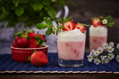 Erdbeer-Panna-Cotta - In einem Glas befindet sich das rosa Erdbeer-Panna-Cotta. Es ist mit Schlagobers, Erdbeeren, einer Blüte und Minze geschmückt. Das Glas steht auf einem blauen Tischtuch. Rechts neben dem Glas sind Kräuterblüten, links ist im Vordergrund ist eine Erdbeere zu sehen und dahinter eine rote, kleine, gefüllte Schüssel. Ein weißes Blumengefäß mit grünen Blumen schmückt den Hintergrund des Bildes. (Foto: VrK/Franz Gleiß - Nicht zur freien Verwendung)