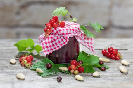 Johannisbeer Chutney - Auf grauem Hintergrund steht ein Einmachglas mit kariertem Tuch über dem Deckel, der Inhalt ist dunkelrot, daneben lieben reife und unreife Johannisbeeren und Blätter. (Foto: VrK/Achim Mandler Photography - Nicht zur freien Verwendung)