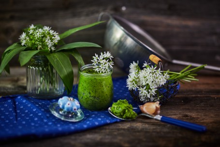 Bärlauchpesto - In der Mitte des Bildes ist ein Glas mit grünem Bärlauch Pesto zu sehen. Dieses ist mit weißen Bärlauch-Blüten verziert. Rundherum befinden sich ein Löffel mit Pesto, ein Körbchen mit Blüten und eine kleine Vase mit Bärlauch-Blättern und Blüten. Das Glas ist mit einem blauen Tischtuch unterlegt. (Foto: VrK/Franz Gleiß - Nicht zur freien Verwendung)