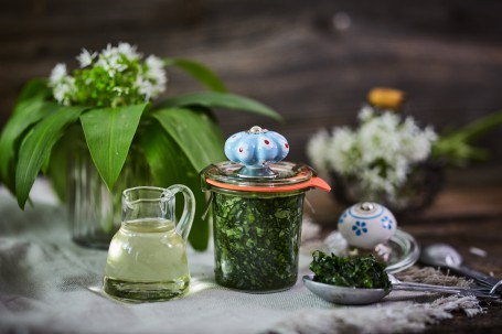 Bärlauch in Öl und Salz - Im Vordergrund befindet sich ein Glas, das mit grünem, eingelegtem Bärlauch gefüllt ist. Auf dem Deckel des Glases ist ein blau-weiß verzierter Griff. Links nebenan steht ein kleiner Krug mit Sonnenblumenöl. Der Hintergrund ist mit einer kleinen Vase mit Bärlauch-Blättern und Blüten versehen. (Foto: VrK/Franz Gleiß - Nicht zur freien Verwendung)