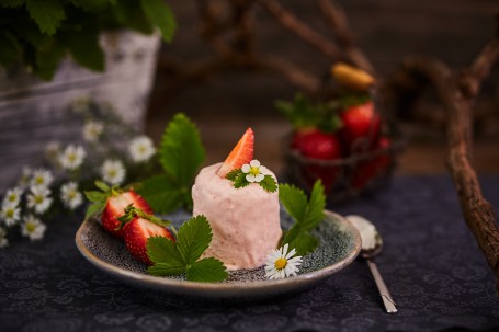 Fruchtparfait - Das Fruchtparfait ist mit Erdbeer-Stücken, -Blättern, und -Blüten und einem Gänseblümchen versehen. Rechts neben dem Teller liegt ein Esslöffel. Im Hintergrund sind Blüten und weitere Erdbeeren zu sehen. (Foto: VrK/Franz Gleiß - Nicht zur freien Verwendung)