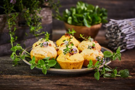 Mais-Speck-Muffins - Auf einem blau-grauen Teller befinden sich sieben Mais-Speck-Muffins. Sie sind mit rosa blühenden Kräutern und grünen Blättern dekoriert. Im Hintergrund ist ein kleines Bündel Holz, ein Blumentopf und eine Blumenkiste mit Kräutern zu sehen. (Foto: VrK/Franz Gleiß - Nicht zur freien Verwendung)