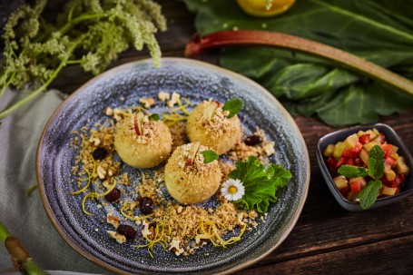 Süße Erdäpfelknödel - Auf einem blau-melierten Teller liegen drei  Erdäpfelknödel. Jeder ist mit einem Rhabarber-Stück bestückt. Der Teller ist zusätzlich mit Rosinen und Streusel  verziert. Im Hintergrund liegen Rhabarber-Blätter und Blüten. (Foto: VrK/Franz Gleiß - Nicht zur freien Verwendung)