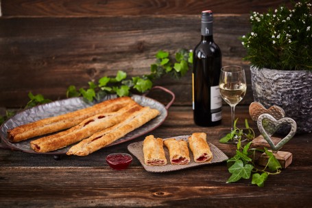 Schmerstrudel - Im Vordergrund sind drei kleine, gold-braune Stück Schmerstrudel auf einem kleinen Teller serviert. Links daneben ist eine kleine Schale mit roter Ribiselmarmelade. Dahinter befindet sich eine Platte mit drei größeren Stücken des Strudels. Im rechten Teil des Bildes ist ein Glas mit Weißwein und dahinter eine Flasche zu sehen. Efeu, kleine Herz-Skulpturen und ein Blumenstock schmücken die Strudelstücke. (Foto: VrK/Franz Gleiß - Nicht zur Verwendung)