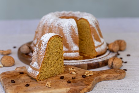 Kaffee-Nuss-Gugelhupf - Auf einem Holzbrett mit Haltegriff ist im Vordergrund ein Stück Gugelhupf zu sehen. Er hat eine gelb-bräunliche Farbe und ist mit Staubzucker bestreut. Vor den Kuchenstück liegen Nussstückchen und Kaffeebohnen zur Deko. Im Hintergrund ist der Rest des Gugelhupfes zu sehen, er steht ebenfalls auf einem Holzbrett und ist mit Staubzucker bestreut. Rund um den Kuchen sind einige Nüsse mit Schale und Kaffeebohnen aufgelegt. (Foto: VrK/Achim Mandler Photography - Nicht zur freien Verwendung)