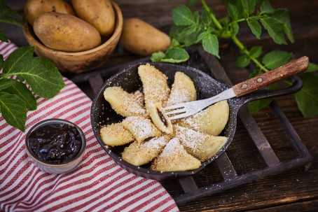 Powidltascherl - Neun Stück Powidltascherl sind in einer schwarzen Pfanne hergerichtet. Beim angeschnittenen Stück liegt eine Gabel. Die Tascherl werden mit Staubzucker bestreut. Links neben der Pfanne ist eine kleine Schüssel mit Powidl. Im Hintergrund sind eine Holzschüssel mit Erdäpfeln und grüne Pflanzen zu sehen. (Foto: VrK/Franz Gleiß - Nicht zur freien Verwendung)