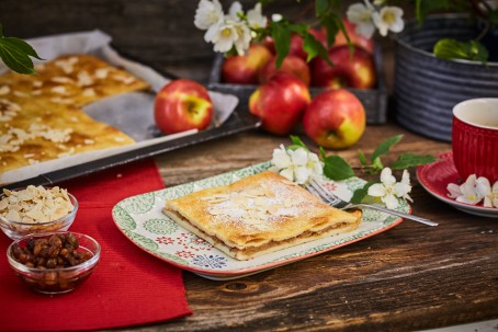 Mürber Apfelkuchen - Der Apfelkuchen ist mit Mandelblättchen und Staubzucker bestreut. Eine kleine Kuchengabel liegt bereit, weiße Blüten und grüne Blätter verzieren den Kuchen. Der Kuchen liegt auf einem weißen Teller mit buntem Muster, links daneben befinden sich zwei kleine Schalen mit Haselnüssen und Mandelblättern. Im Hintergrund ist das Backblech mit dem Kuchen zu sehen, rechts daneben steht eine Obstschüssel mit Äpfeln. (Foto: VrK/Franz Gleiß - Nicht zur freien Verwendung)