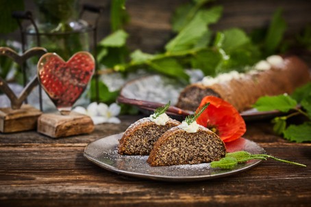 Mohnkuchen - Auf einem dunkelbraunen Holztisch und einem grau-braunen Teller werden zwei Stück Mohnkuchen serviert. Auf dem Teller befinden sich zur Zierde grüne Blätter und eine Mohnblüte. Im Hintergrund sieht man den Mohnstrudel, zwei kleine, herzförmige Skulpturen und grüne Blätter. (Foto: VrK/Franz Gleiß - Nicht zur freien Verwendung)