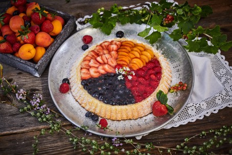 Früchtekuchen - Der Früchtekuchen wird auf einem silbernen Teller serviert. Die Oberfläche des Kuchens bilden Heidelbeeren, Himbeeren, Erdbeeren und Marillen. Zusätzlich ist der Teller mit Ribiseln geschmückt. Links im Hintergrund befindet sich eine Obstschale mit Früchten. Der Teller ist mit einem weißen Tischtuch mit Spitzen unterlegt. (Foto: VrK/Franz Gleiß - Nicht zur freien Verwendung)