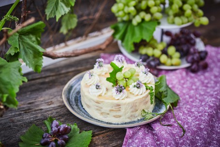 Weincremetorte - Die Weitcremetorte steht auf einem blau-melierten Teller. Dieser befindet sich auf einem  dunkelbraunen Holztisch. Der Teller ist mit einem violetten Tischtuch mit weißen Verzierungen unterlegt. Auf der Weincremetorte sind 8 Kügelchen aus Schlagobers, die mit violetten Blüten bedeckt sind. In der Mitte befinden sich grüne Weintrauben mit einem grünen Blatt. Im Vordergrund des Bildes sind dunkelblaue Weintrauben, die auf grünen Blättern liegen. Im Hintergrund befinden sich Weinpflanzen mit einem Holzspalier. (Foto: VrK/Franz Gleiß - Nicht zur freien Verwendung)