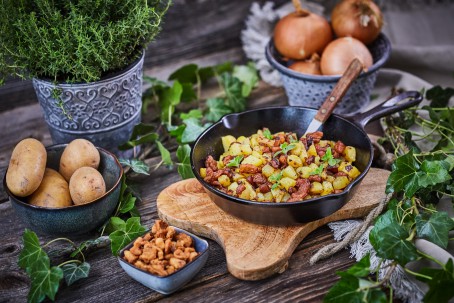 Grammelsterz - Grammelsterz wird in einer schwarzen Pfanne auf einem Holzuntersetzer serviert. In einer kleinen, blauen Pfanne befinden sich Grammel. Links nebenan sind in einer Schüssel  vier Stück Erdäpfel platziert. Im Hintergrund befinden sich eine blaue Schüssel mit Zwiebel und ein Kräutertopf mit Thymian. (Foto: VrK/Franz Gleiß - Nicht zur freien Verwendung)