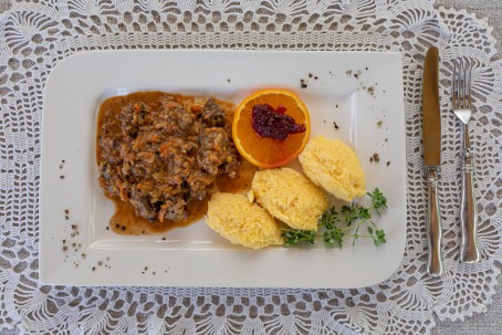 Hirschragout mit Polentanockerl - Auf einem weißen Spitzendeckchen steht ein an zwei Ecken abgerundeter weißer Teller. Auf dem Teller liegen in der rechten vorderen Ecke 3 gelbe Polentanockerl. Daneben liegen einige Halme grüner Kräuter. In der linken Hälfte des Tellers ist das bräunliche Hirschragout platziert. Die Fleischstücke und auch Karottenstücke sind gut erkennbar. Rechts neben dem Ragout, oberhalb der Polentanockerl kann man eine Orangenscheibe mit Preiselbeermarmelade sehen. Rechts neben den Teller liegen Messer und Gabel schon bereit. (Foto: VrK/Achim Mandler Photography - Nicht zur freien Verwendung)