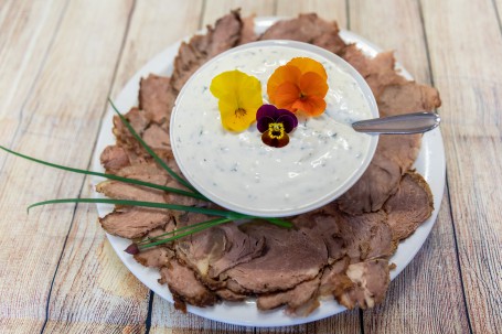 Roastbeef mit Kräutertatar - Auf einem hellen Holzhintergrund steht ein weißer runder Teller. AUf dem Teller liegen fein aufgeschnittene Roastbeefscheiben. In der Mitte steht eine weiße Schüssel mit der weißen Kräutertatar, die Kräuter sind zu erkennen. Auf der Kräutertatar liegen dekorativ 3 Blüten eine gelbe, eine orange und eine lila farbige. In der Kräutersoße steckt ein silberner Löffel. (Foto: VrK/Achim Mandler Photography - Nicht zur freien Verwendung)