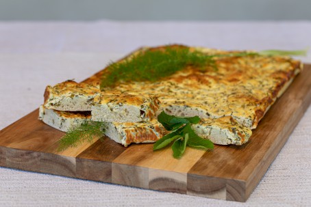 Warmer Kräuterauflauf - Auf einem Holzbrett liegt ein wie Pizzateig aussehender Teig. Der Auflauf ist rechteckig und hat eine gelbe Oberseite. Die Kräuter sind gut erkennbar. Garniert ist das ganze mit Dill (Foto: VrK/Achim Mandler Photography - Nicht zur freien Verwendung)