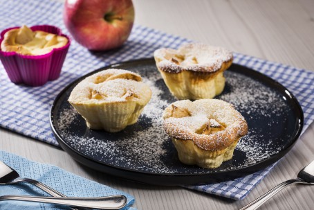 Apfelküchlein - Auf einem schwarzen runden Teller sind 3 Muffinförmige Küchlein angerichtet. Die Apfelstückchen sind deutlich zu erkennen. Die Küchlein sind mir Staubzucker bestreut. Im Hintergrund steht eine rote Muffinform mit einem weiteren Muffin darin. Ebenso im Hintergrund liegt ein roter Apfel. (Foto: VrK/Alexander Stiegler - Nicht zur freien Verwendung)