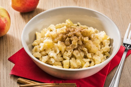 Apfelnockerl - Die wie Spätzle aussehenden Apfelnockerl sind in einer weißen Suppenschüssel angerichtet und mit Zimtpulver bestreut. Die Schüssel steht auf einer dunkelroten Serviette. Rechts neben der Schüssel liegt eine Speisegabel. Als Deko liegen im Hintergrund 2 Äpfel und im Vordergrund 2 Zimtstangen. (Foto: VrK/Alexander Stiegler - Nicht zur freien Verwendung)