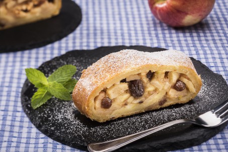 Apfelstrudel mit Topfenteig - Auf einem blau-weiß-karriertem Tischtuch steht eine schwarze runde Schieferplatte. Darauf befindet sich ein Stück Apfelstrudel. Die Apfelstücke und Rosinen sind deutlich zu erkennen. (Foto: VrK/Alexander Stiegler - Nicht zur freien Verwendung)